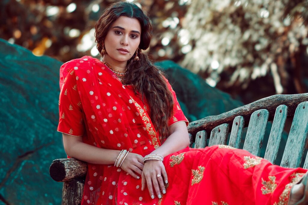 A woman in a vibrant red saree sits elegantly on a wooden bench outdoors.