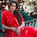 A woman in a vibrant red saree sits elegantly on a wooden bench outdoors.