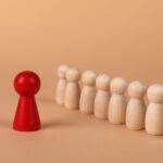 A red pawn stands out as it leads a group of wooden pawns on a beige background symbolizing leadership.