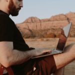 Side view of crop male freelancer sitting on chair while working on laptop in desert with mountain range