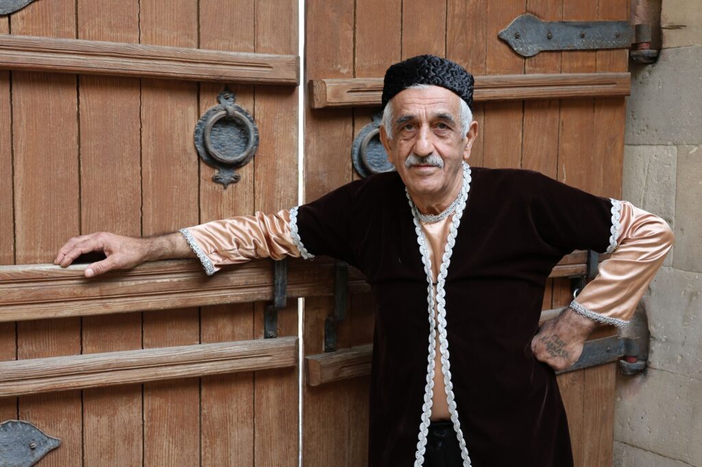 Senior man in traditional clothing stands by rustic wooden doors.