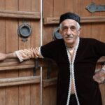 Senior man in traditional clothing stands by rustic wooden doors.