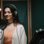 Woman singing into a microphone in a professional music studio wearing headphones.