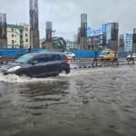 Cars navigate a flooded street in Kolkata, India, amidst heavy rain and overcast skies.