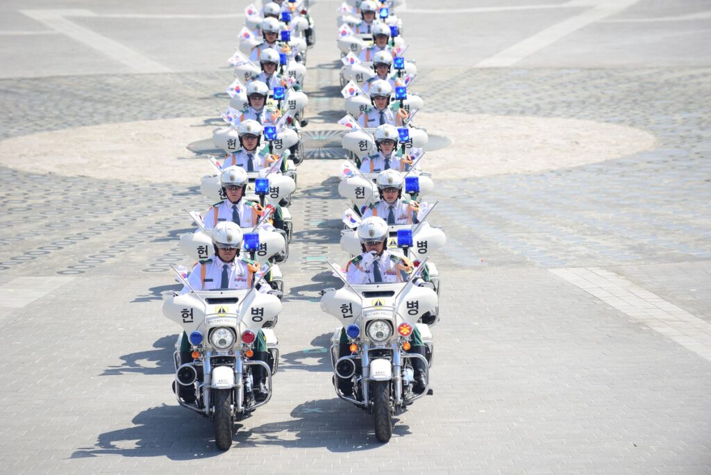 A line of South Korean police officers on motorcycles holding flags in an urban parade.
