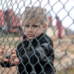 Sad ethnic child standing near metal net against freight car and looking at camera