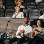 A couple enjoys popcorn while watching a movie in a cozy cinema environment, surrounded by others.