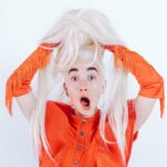 A startled woman in a vibrant orange outfit holding a blonde wig with a surprised expression.