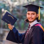 Smiling graduate in cap and gown holds diploma outdoors, celebrating academic achievement.