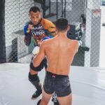 Two athletes engaged in a mixed martial arts sparring session inside a cage.