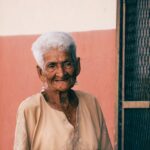 An elderly woman with white hair smiles warmly in front of a colorful wall.