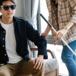 A visually impaired man receives support from a caregiver indoors, using a walking stick.