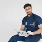 Young male doctor in blue scrubs reviewing medical records with a confident smile.