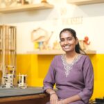 A smiling woman in a purple dress in a DIY workshop setting.