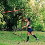Athletic woman practicing javelin throw in a vibrant green forest setting during daytime.