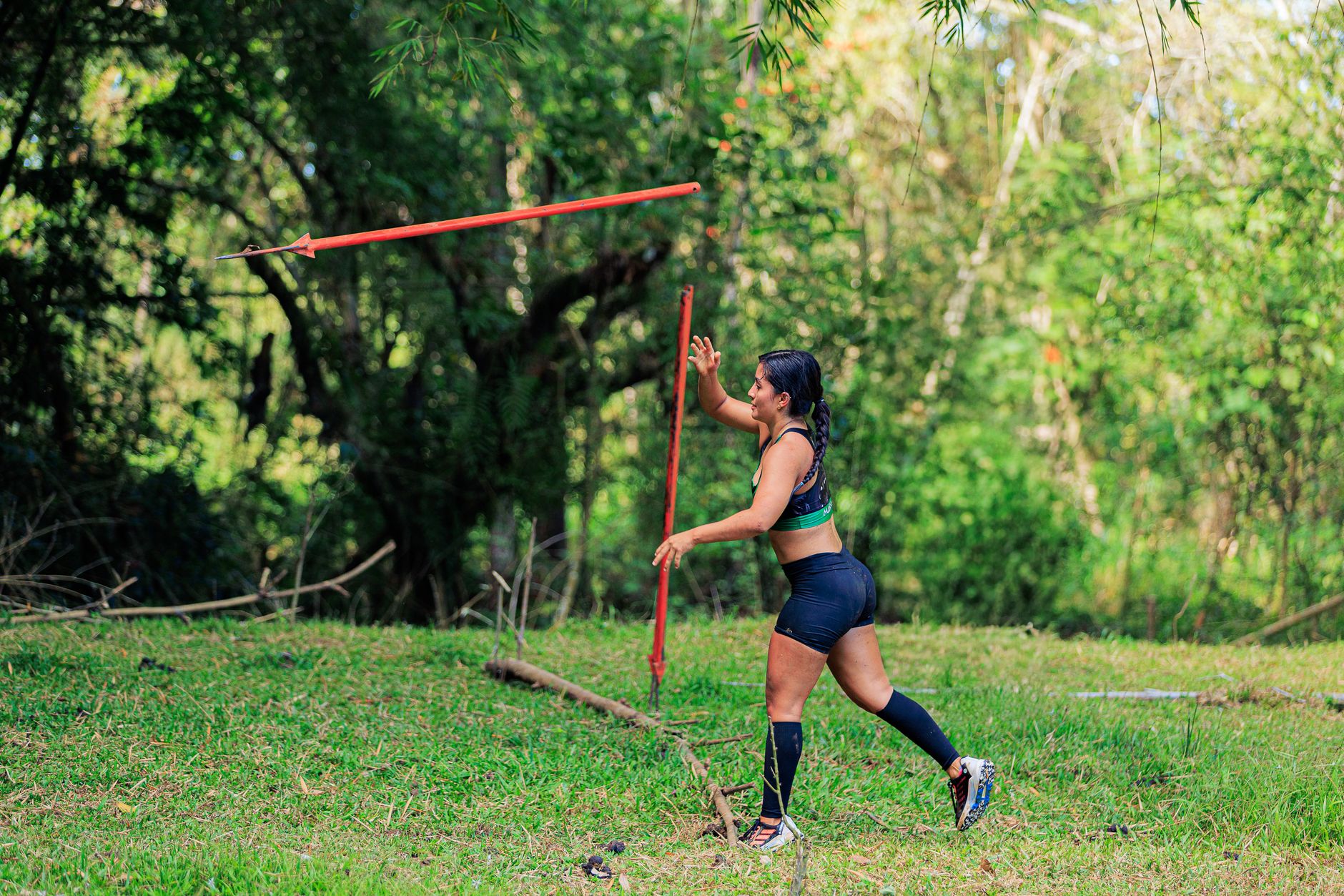 Athletic woman practicing javelin throw in a vibrant green forest setting during daytime.
