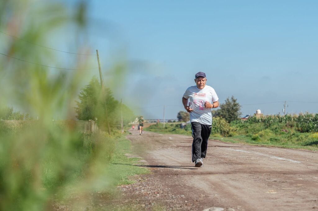 Elderly man jogging on a rural dirt road, wearing casual sportswear in sunny weather.