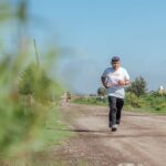 Elderly man jogging on a rural dirt road, wearing casual sportswear in sunny weather.