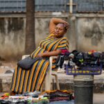 A vendor sits on the sidewalk with merchandise in an urban city setting, selling clothing on a warm day.