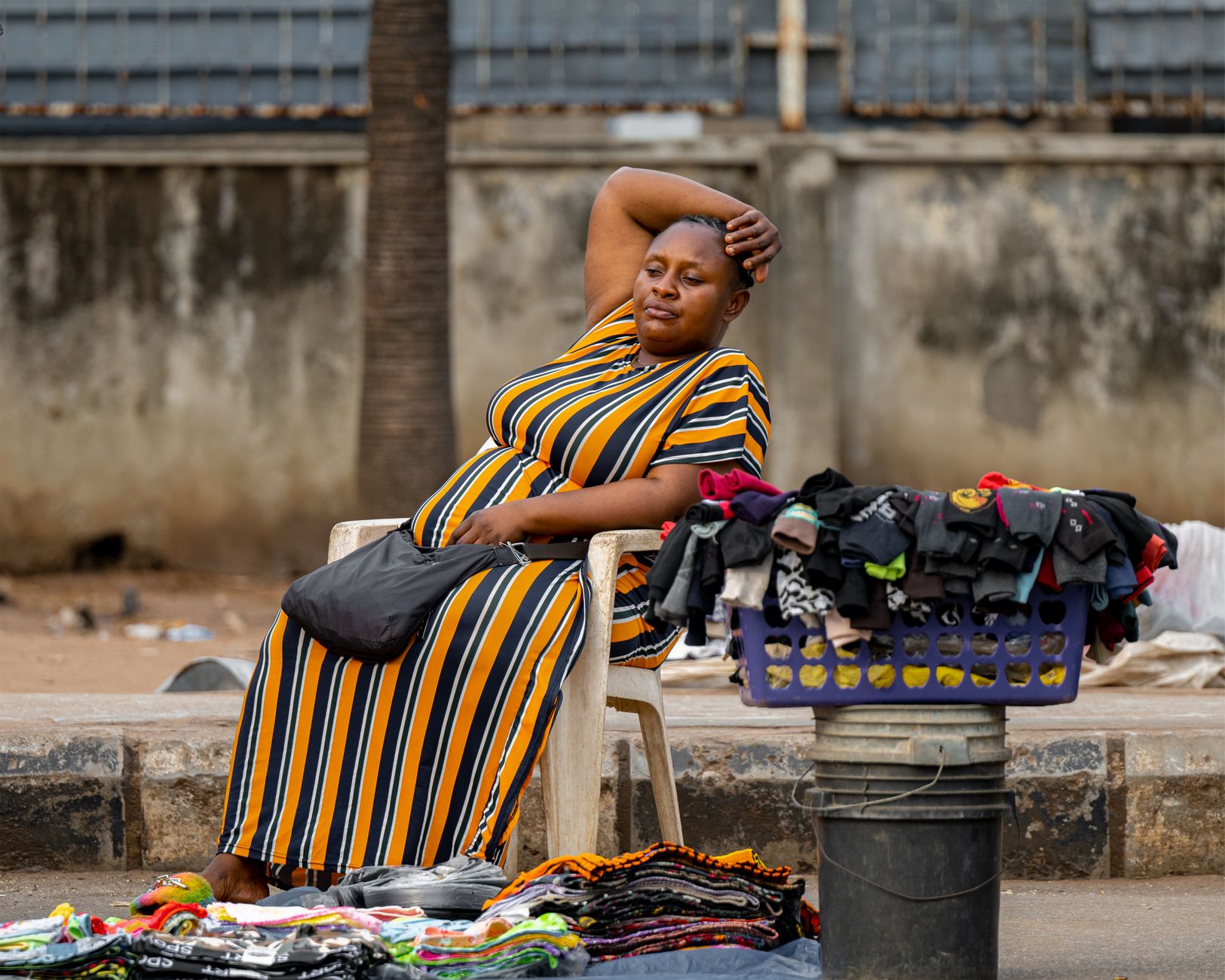 A vendor sits on the sidewalk with merchandise in an urban city setting, selling clothing on a warm day.