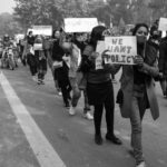 Black and white photo of a protest rally with people holding 'We Want Policy' signs.