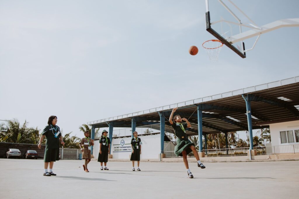 Group of children in school uniforms playing basketball outdoors on a sunny day.