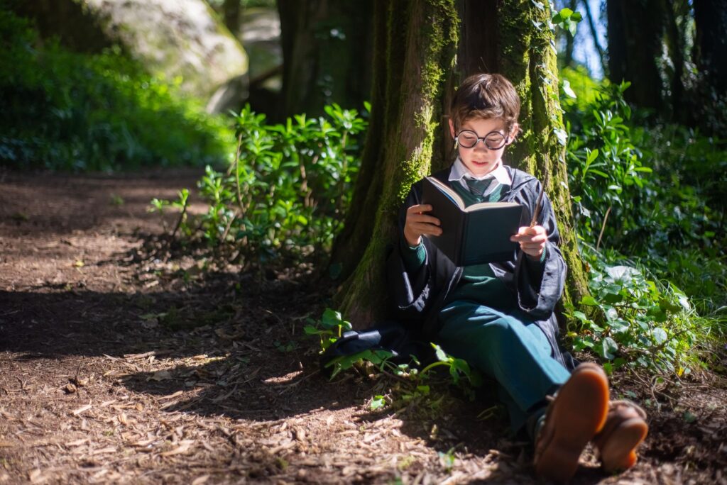 Child in wizard costume reading a book under a tree in a sunlit forest.