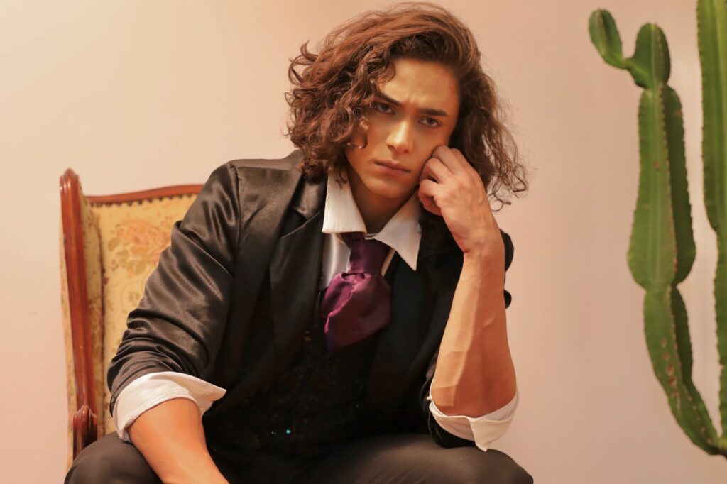 Young man with curly hair in formal attire sitting indoors with cactus in background.