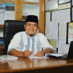 An Indonesian teacher in traditional attire, smiling at his desk with documents in an office setting.