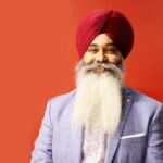 Portrait of a smiling Sikh man with a long beard and turban against a red background.