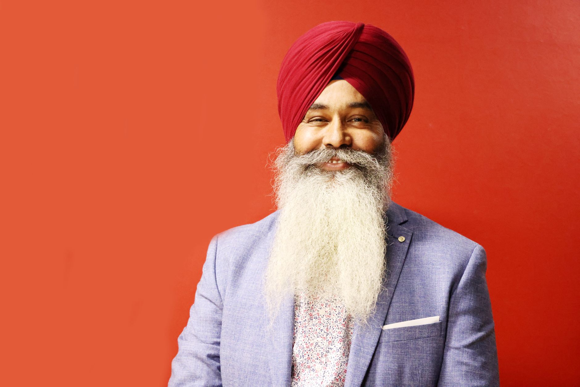 Portrait of a smiling Sikh man with a long beard and turban against a red background.