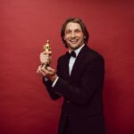 Smiling man in a tuxedo holding a trophy against a red background, celebrating success.