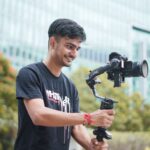 Smiling young videographer using a gimbal outdoors. Urban setting and greenery in the background.