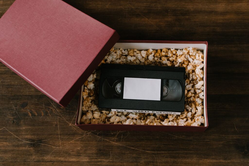An open gift box with a VHS tape nestled in popcorn, on a wooden surface.