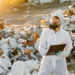 Scientist in protective gear documenting pollution at a garbage dump site outdoors.