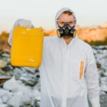 Worker in a protective suit holds a yellow container, monitoring pollution at a waste site.