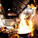 A skilled chef performs a dramatic flambé cooking technique in a professional kitchen setting.