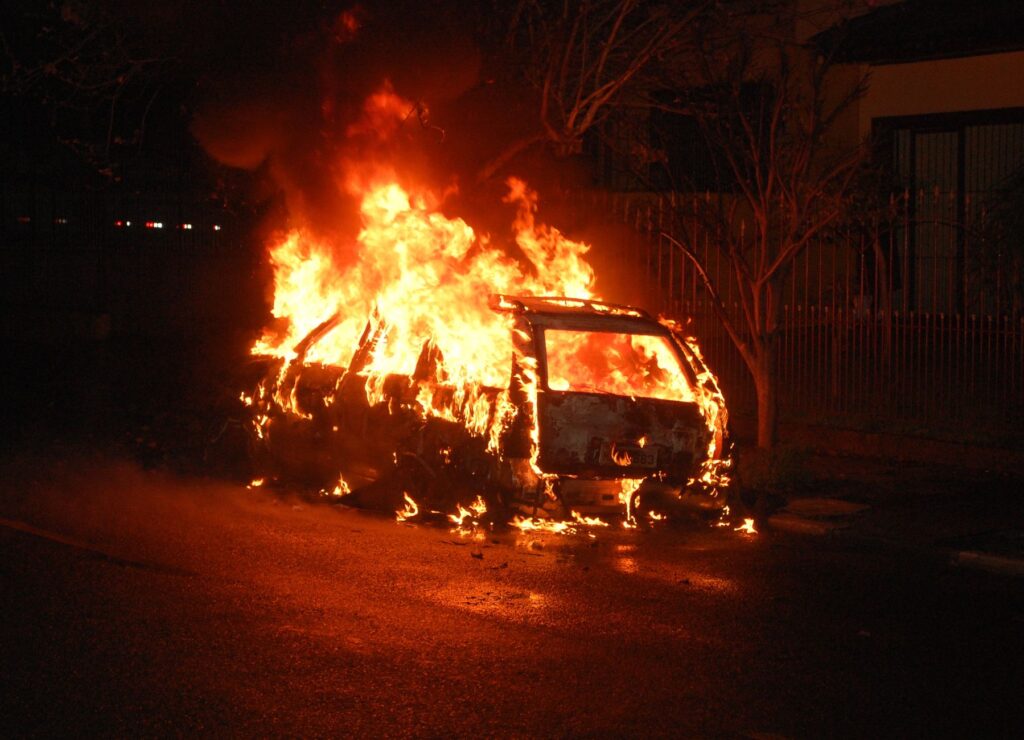 A vehicle is completely engulfed in flames, casting a dramatic glow on a dark street.