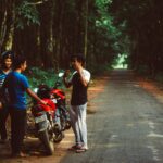 A casual group of friends chatting beside motorbikes on a tranquil forest road in Mainama, India.