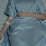 A patient lies in bed wearing a hospital gown with an IV drip attached, evoking a sense of illness.