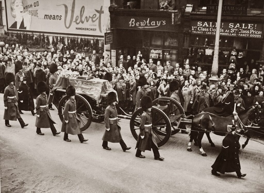 Historical black and white photo of a royal funeral procession with soldiers and crowd in city street.
