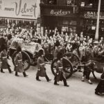 Historical black and white photo of a royal funeral procession with soldiers and crowd in city street.