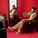 Fashionable young man sitting in front of a mirror against a vivid red backdrop, showcasing style and elegance.