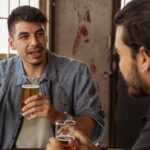 Two men enjoying a casual conversation with beers in an indoor café setting.