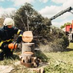 A firefighter uses a chainsaw to cut a tree stump while a truck assists in an outdoor setting.