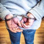 Close-up of a person in handcuffs viewed from behind, wearing casual clothing indoors.