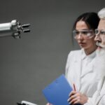 Two scientists wearing lab coats and goggles analyzing a robotic arm in a laboratory setting.