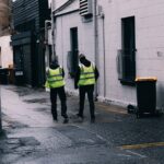 Two safety-vested men walk in a rainy urban alley, next to estate agency.