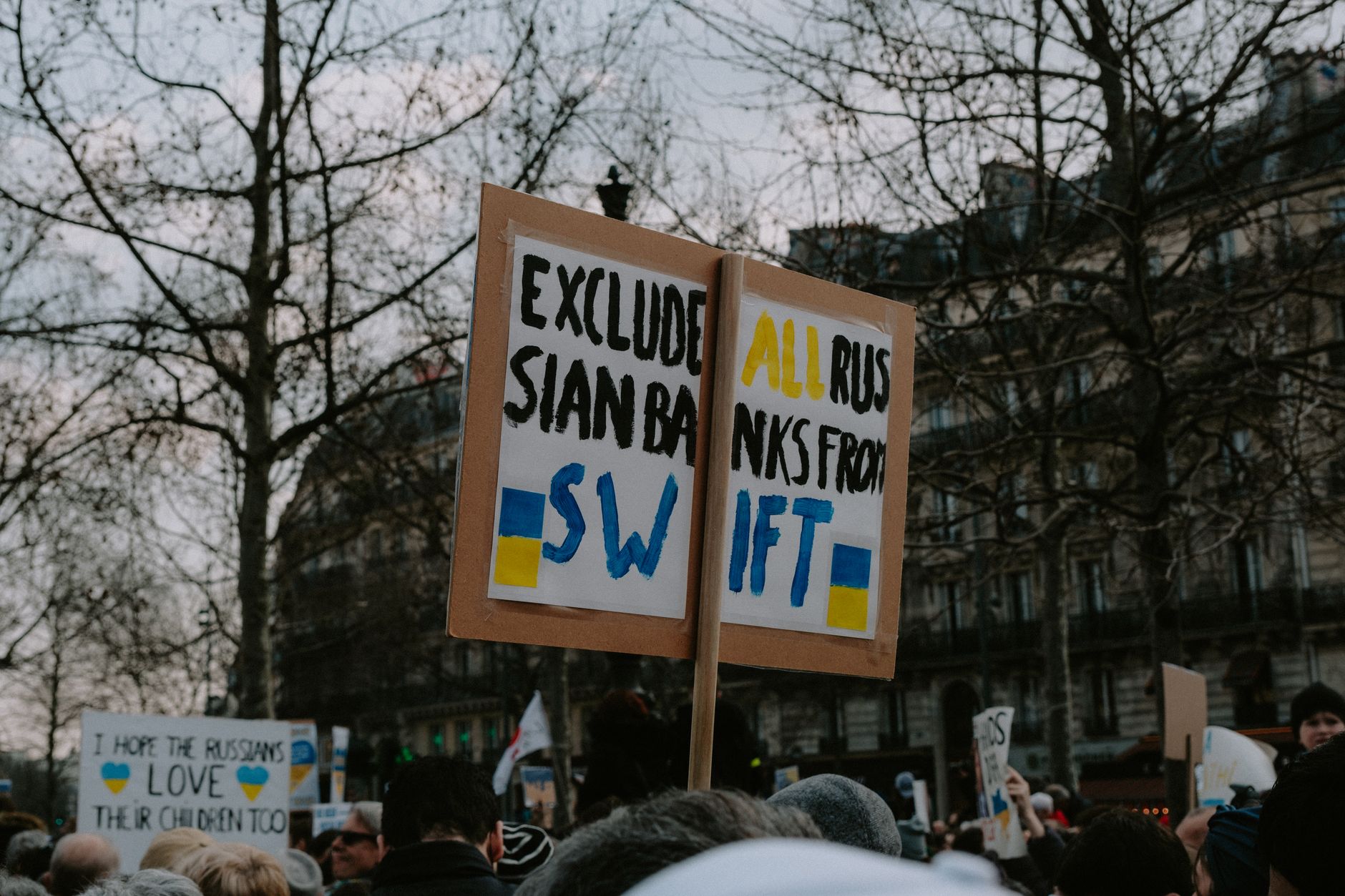 Crowd gathered with placards during a protest against Russian banks in an urban setting.
