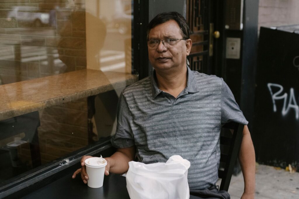 Concentrated Asian male in eyeglasses sitting near window at table with cup of hot drink in outdoor cafe during break